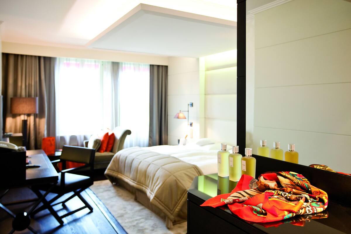 A hotel room with a bed, a desk, a chair, a lamp, a mirror, and a table with bottles and a colorful cloth.