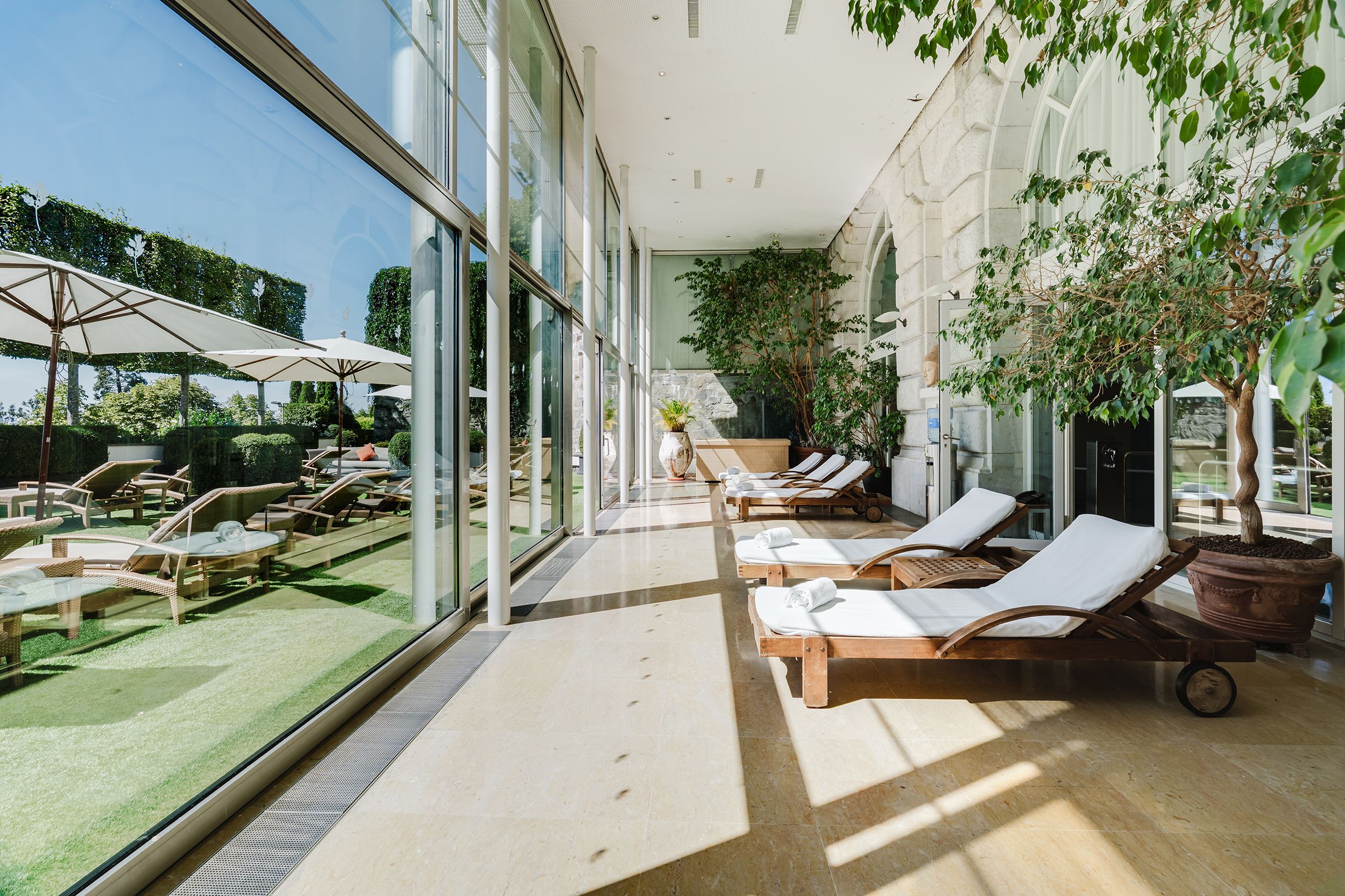 A sunlit indoor patio with lounge chairs and umbrellas, large windows, and indoor plants.