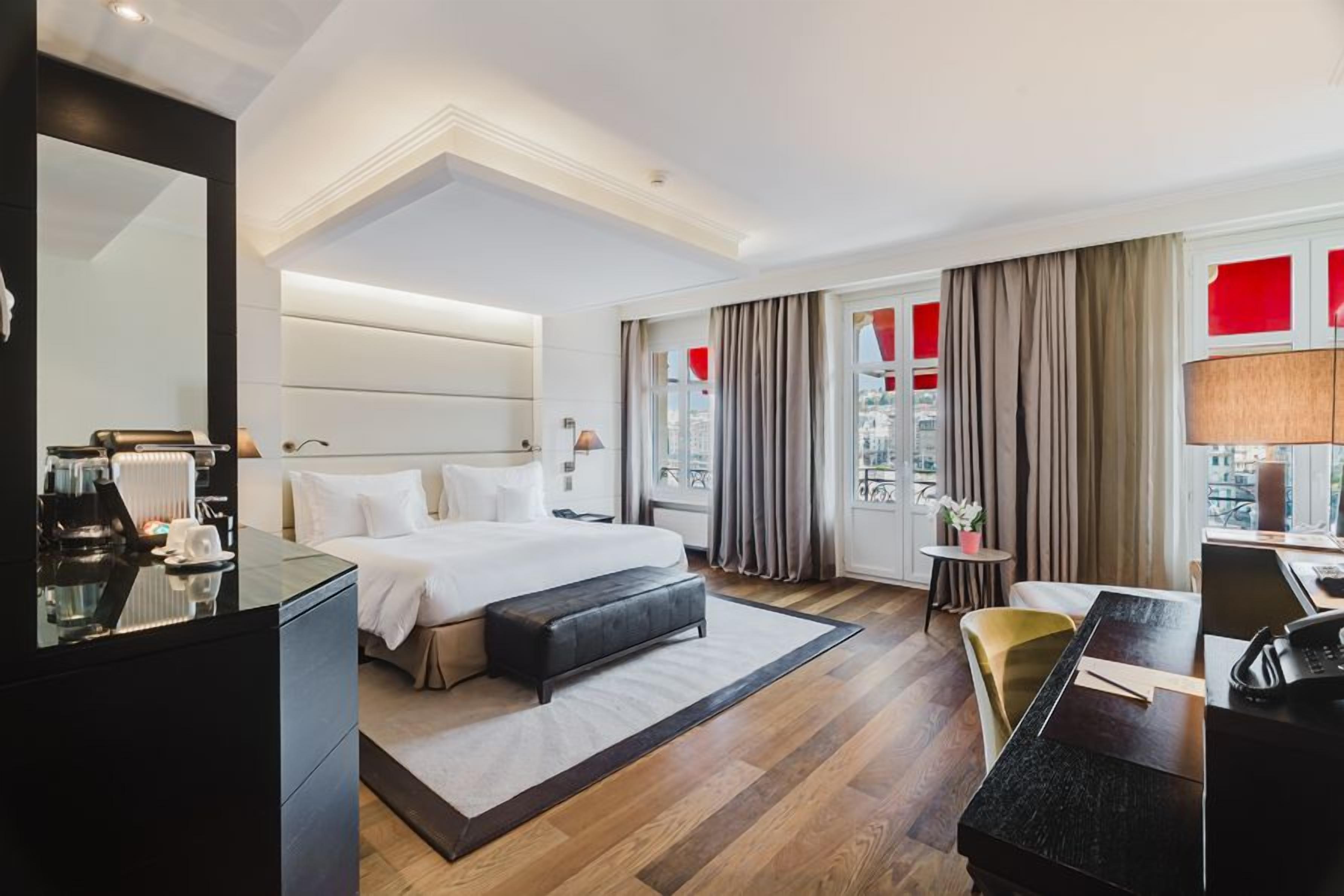 A hotel room with a large bed, a desk, a chair, a mirror, and a view of the city.