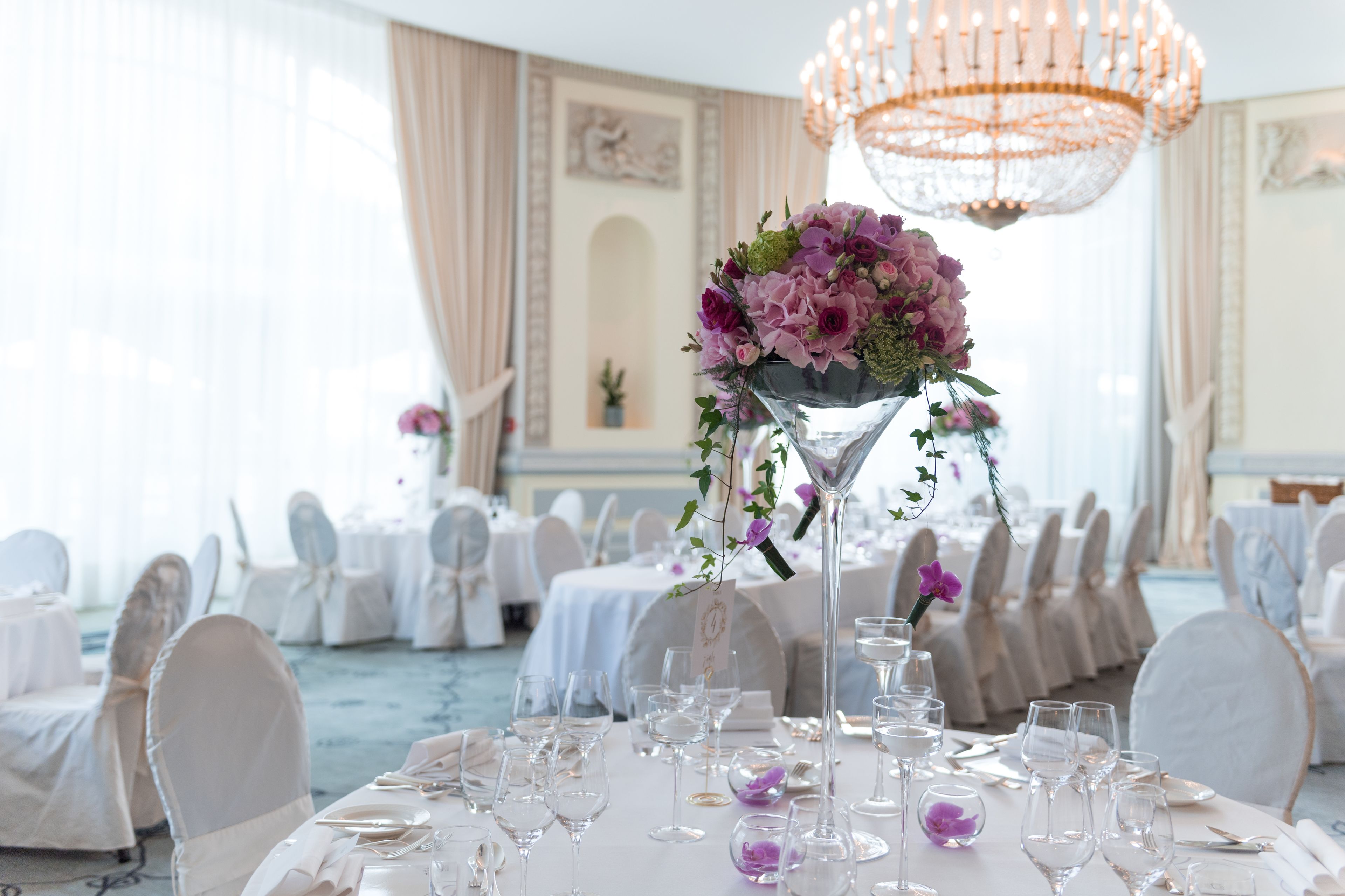 A banquet hall with round tables set for a formal event, featuring a central floral arrangement and elegant lighting.