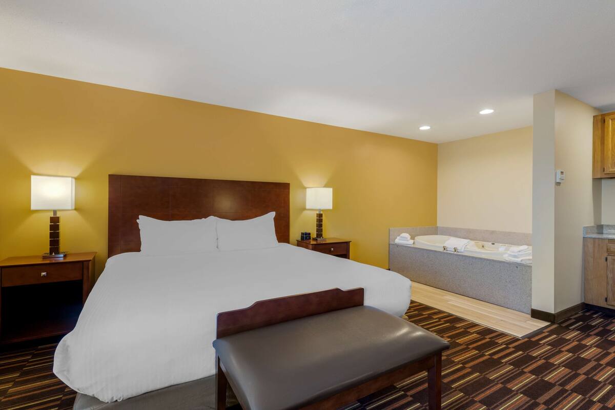 Une chambre d’hôtel avec un grand lit, des lampes de chevet, un banc, une baignoire et une armoire en bois.