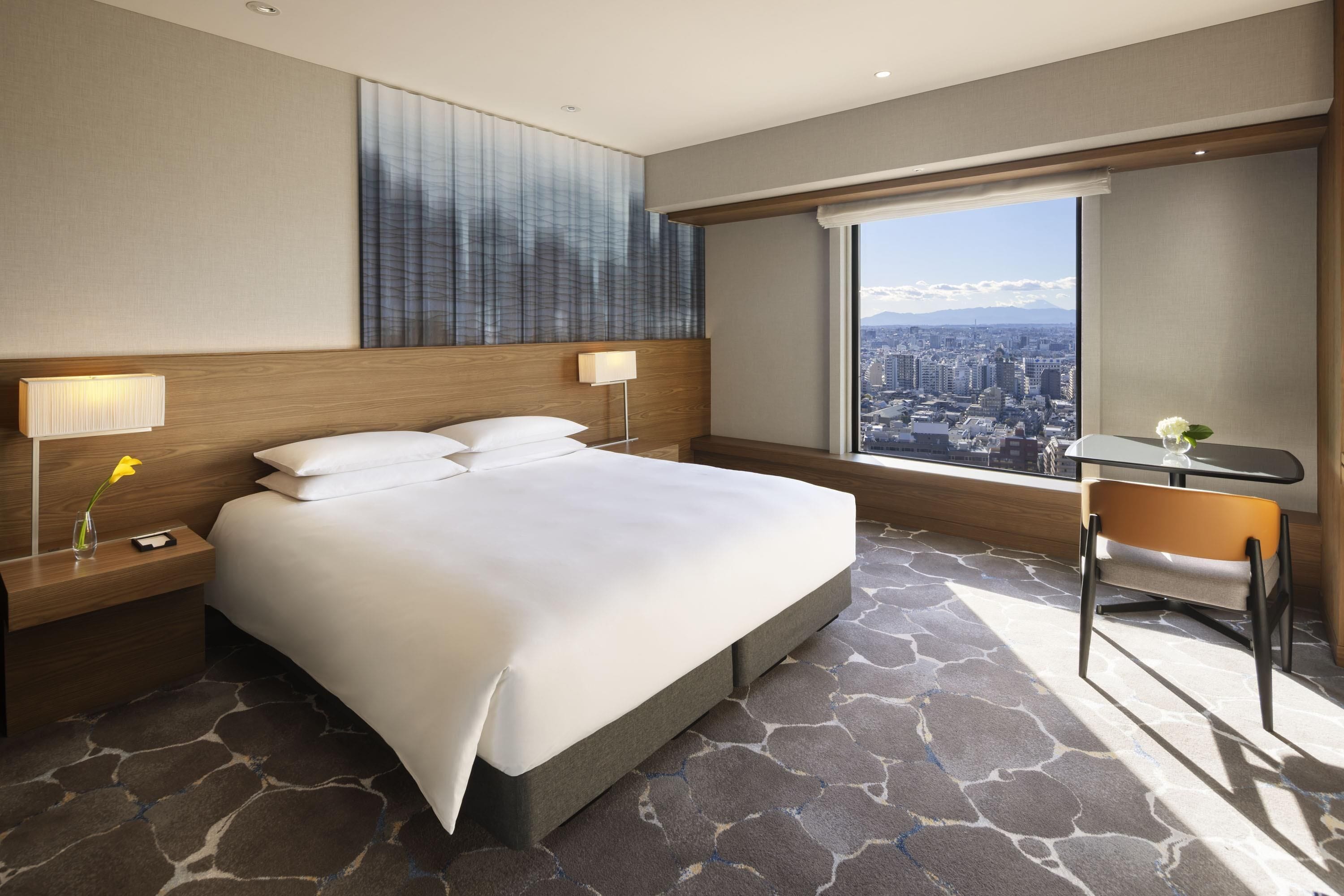 A hotel room with a large bed, two bedside lamps, a nightstand, a chair, a table, and a city view.