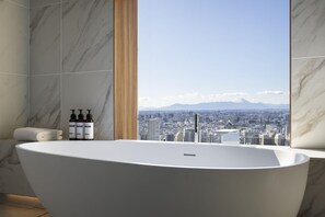 A modern bathroom with a freestanding bathtub, marble walls, a city view, and toiletries on the side.