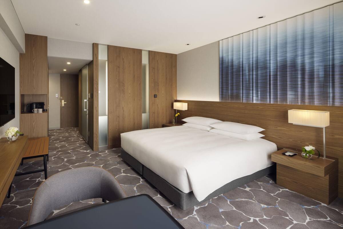 A modern hotel room with a large bed, bedside lamps, a desk with a coffee maker, and a TV.