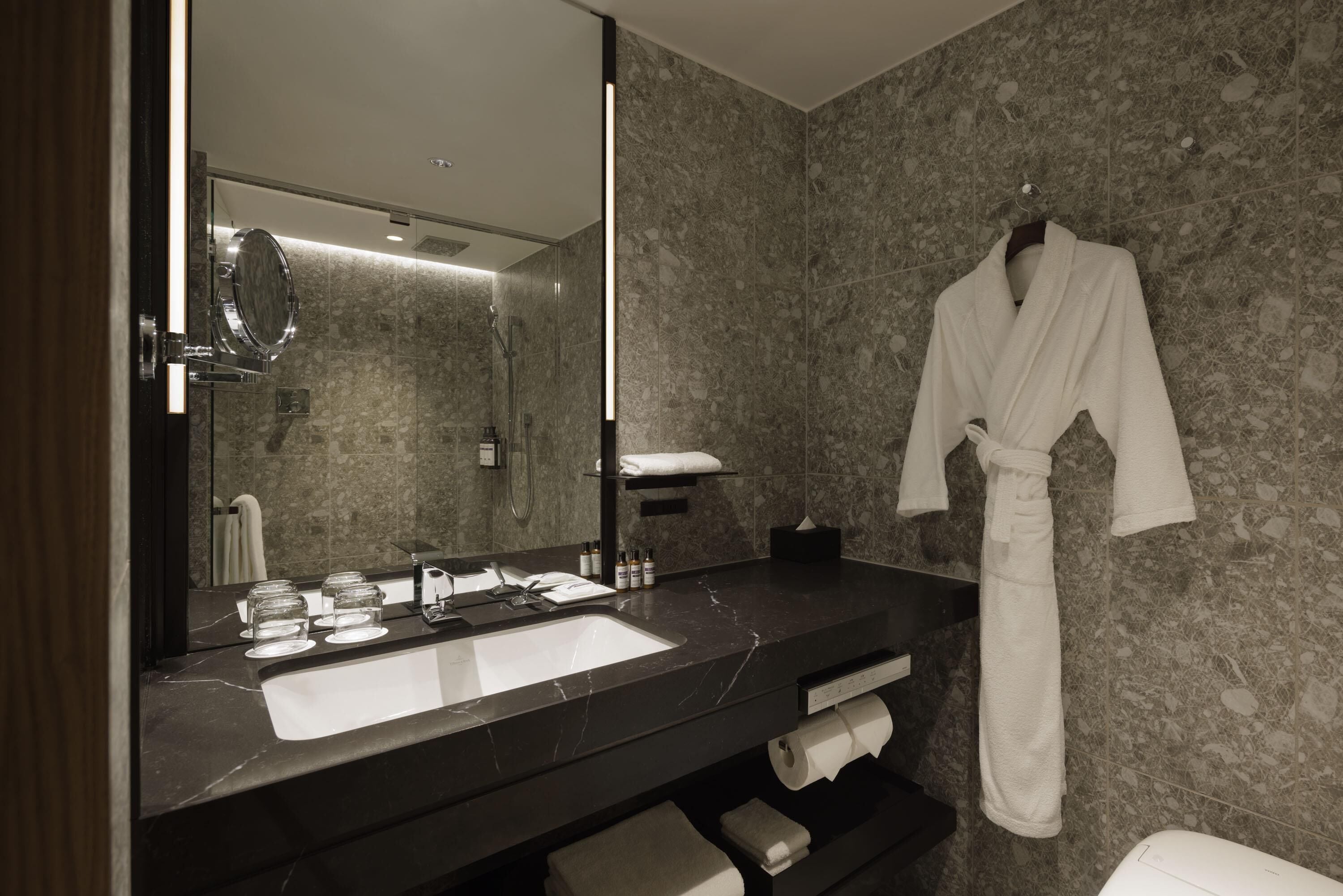 Un baño moderno con un espejo grande, encimera de mármol oscuro y una bata colgada en la pared.