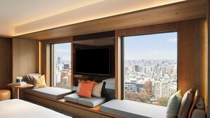 A modern hotel room with a large window, a flat-screen TV, a sofa bed, and a small table with a lamp.