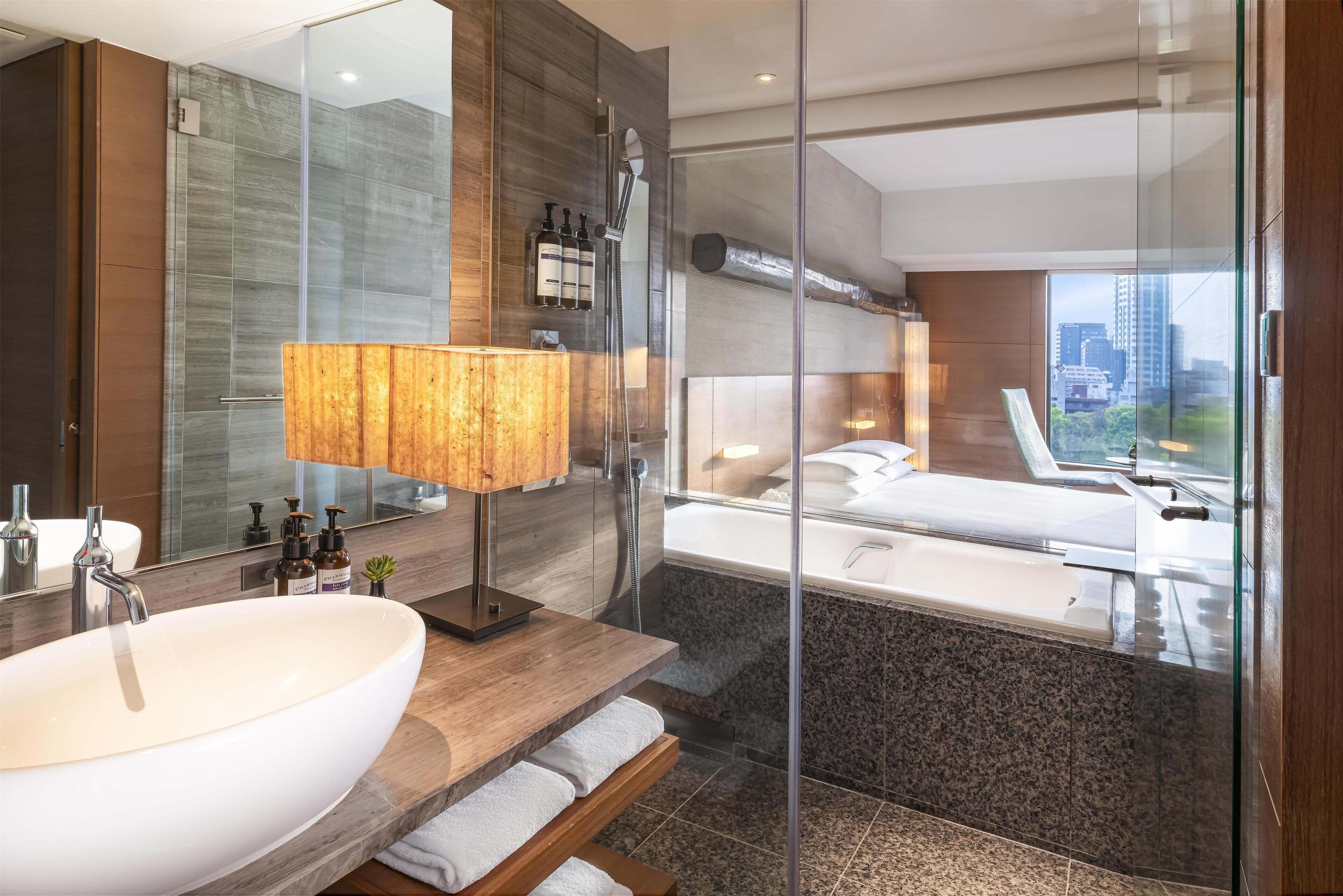 A modern bathroom with a large white sink, a glass shower enclosure, and a view of a cityscape through a large window.