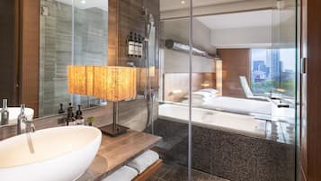 A modern bathroom with a large white sink, a glass shower enclosure, and a view of a cityscape through a large window.