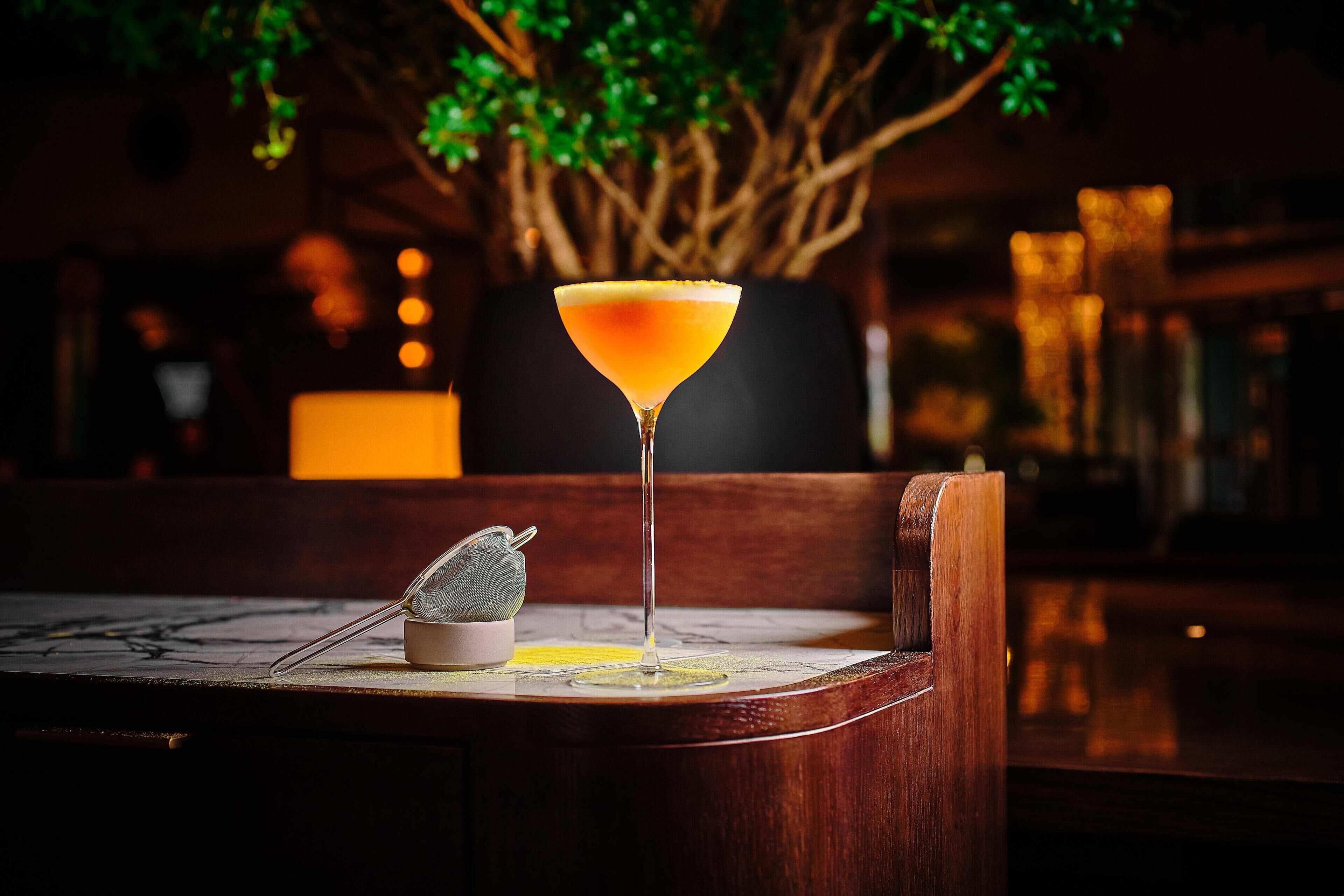 Ein Martini-Glas auf einem Holztisch, zusammen mit einem Barsieb, einer Kerze und einem Baum im Hintergrund.