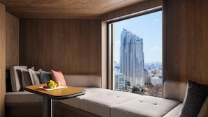 A modern hotel room with a large window offering a city view, a sofa with cushions, a small table with fruits, and a patterned pillow.