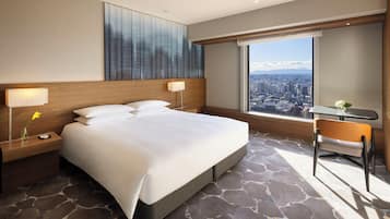 A hotel room with a large bed, two bedside tables with lamps, a small table with a chair, and a large window offering a city view.