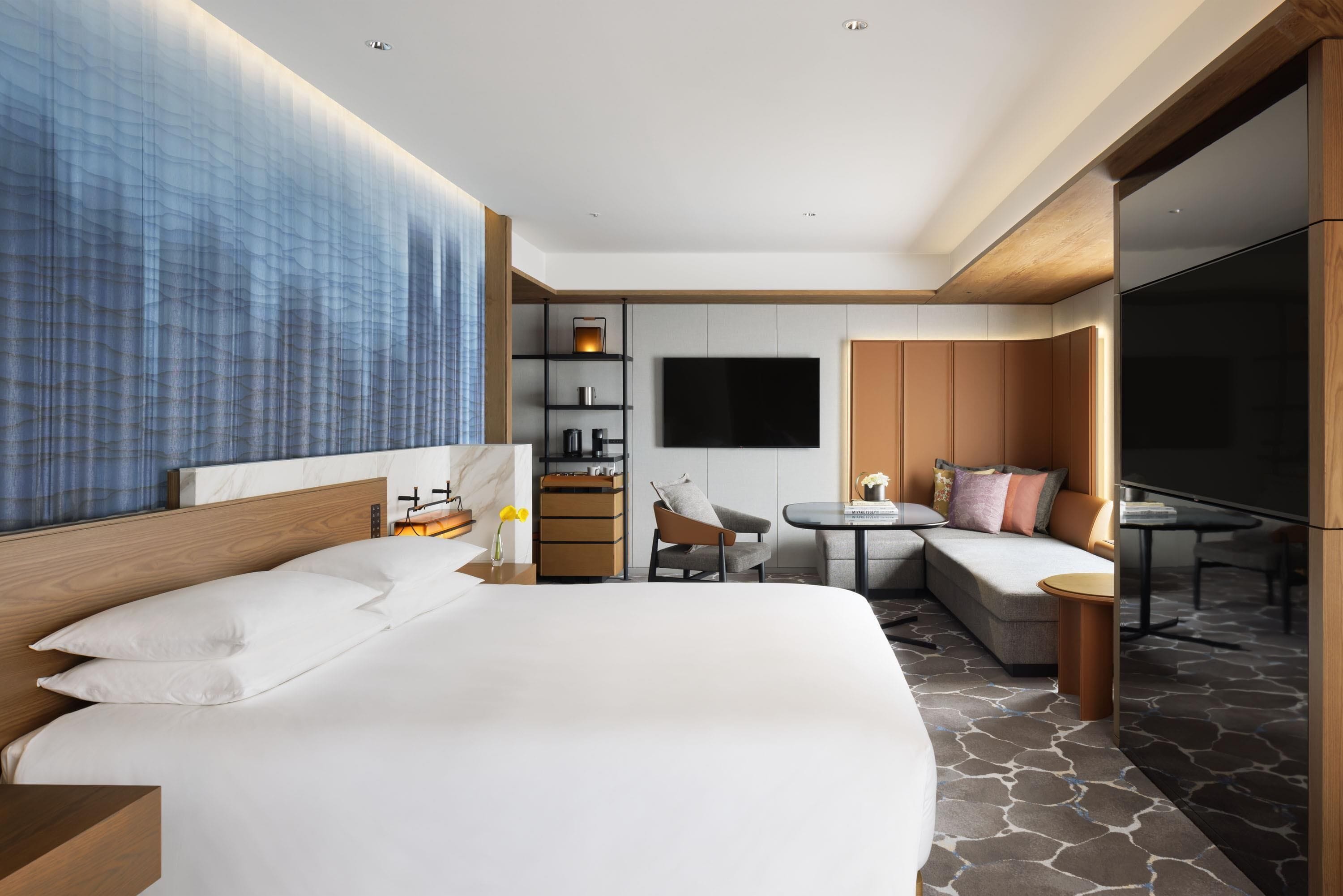 A modern hotel room with a large bed, a TV, a seating area with a sofa and chairs, a small table, and a wardrobe.