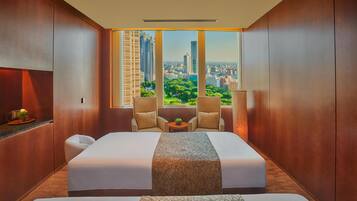 Habitación de hotel con una cama grande, dos sillones, una mesita y vistas a un paisaje urbano a través de amplios ventanales.