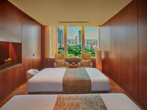 A hotel room with a large bed, two armchairs, a small table, and a view of a cityscape through large windows.