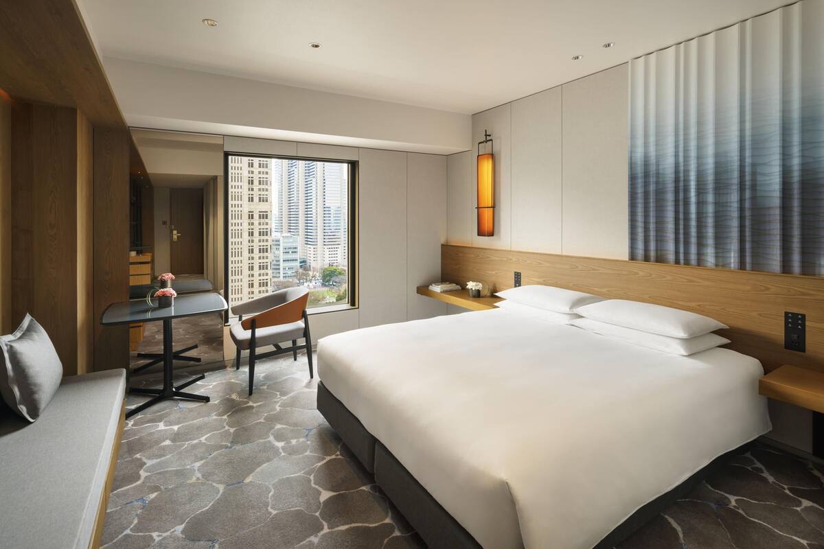 A hotel room with a large bed, a desk, a chair, a sofa, a window with a city view, and a wall-mounted lamp.