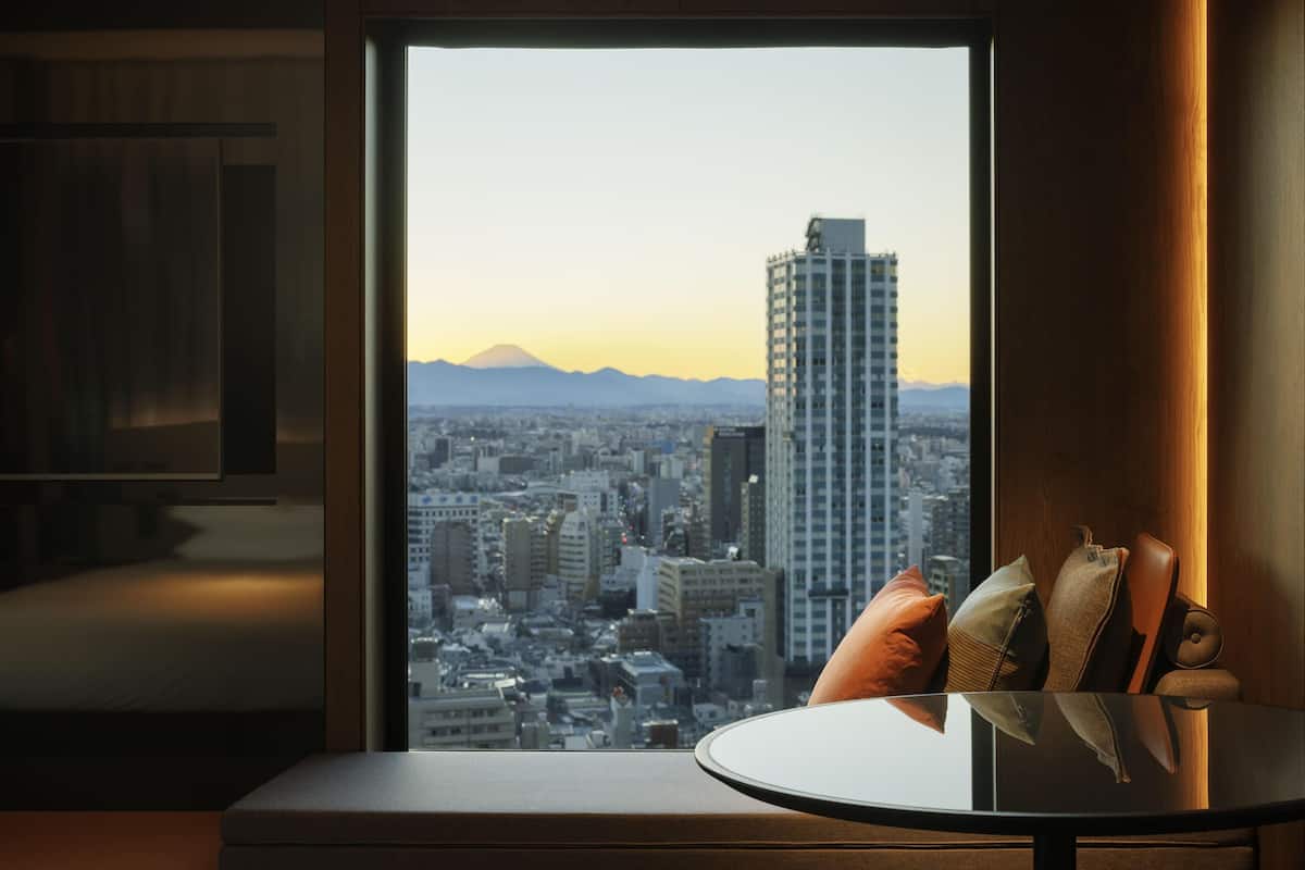 A room with a large window overlooking a cityscape, a round table with cushions, and a wooden floor.