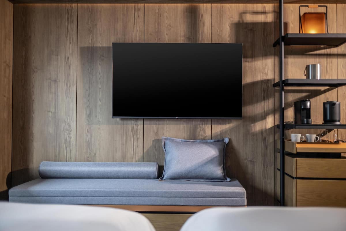 A modern room with a flat-screen TV mounted on a wooden wall, a grey sofa with cushions, and a wooden cabinet with shelves.
