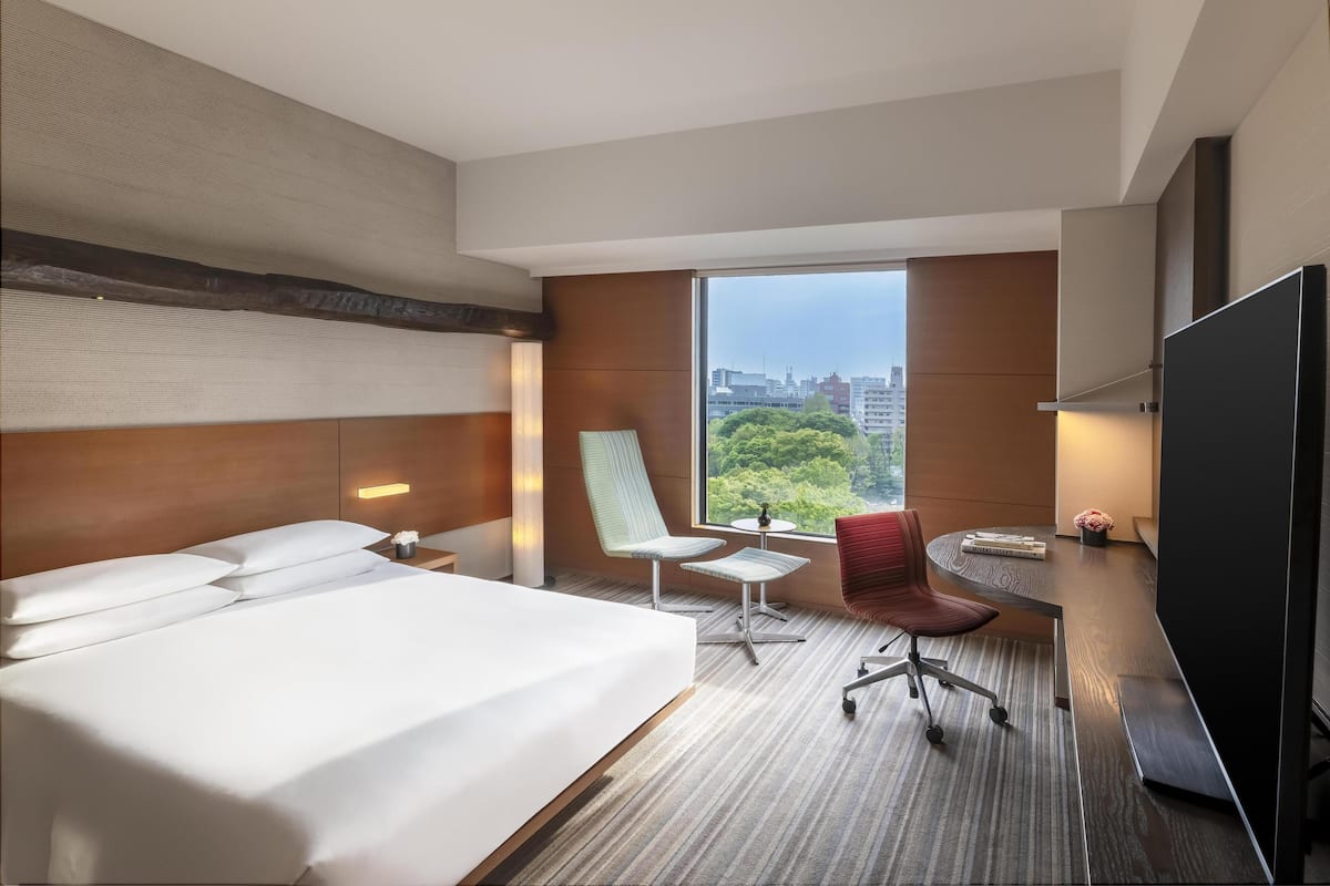 A hotel room with a large bed, two chairs, a small table, a TV, and a large window with a city view.