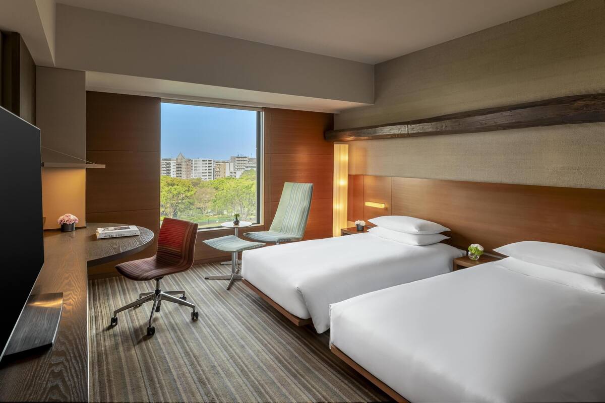 A hotel room with two beds, a desk, a chair, a TV, and a large window with a city view.