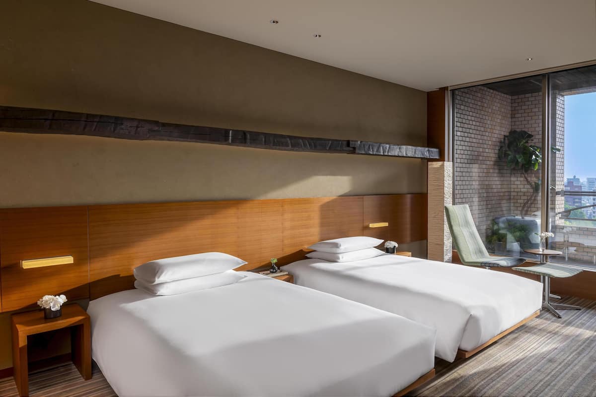 A hotel room with two beds, a wooden headboard, a small table with a chair, and a large window with a view.