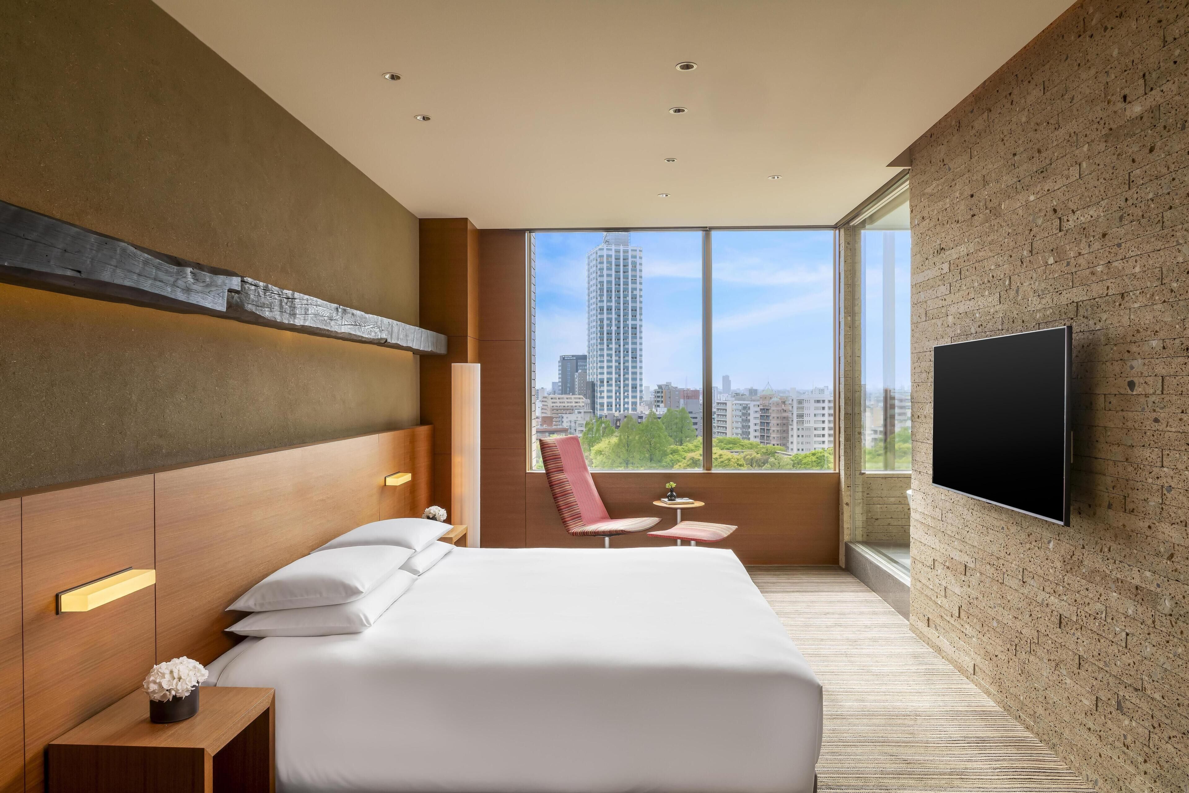 Una habitación de hotel moderna con una cama grande, un televisor montado en la pared, vistas a la ciudad a través de amplios ventanales y una pequeña zona de estar.