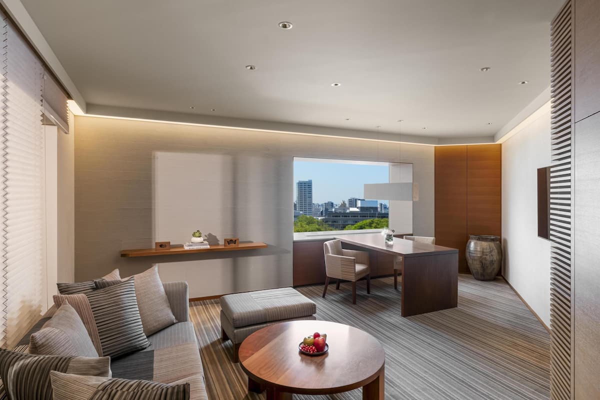 A modern hotel room with a sofa, a round table with fruit, a desk, and a large window offering a city view.