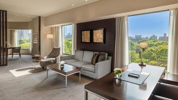 Habitación de hotel moderna con sofá, sillón, mesa de centro y escritorio con lámpara. Amplias ventanas ofrecen vistas a la ciudad.