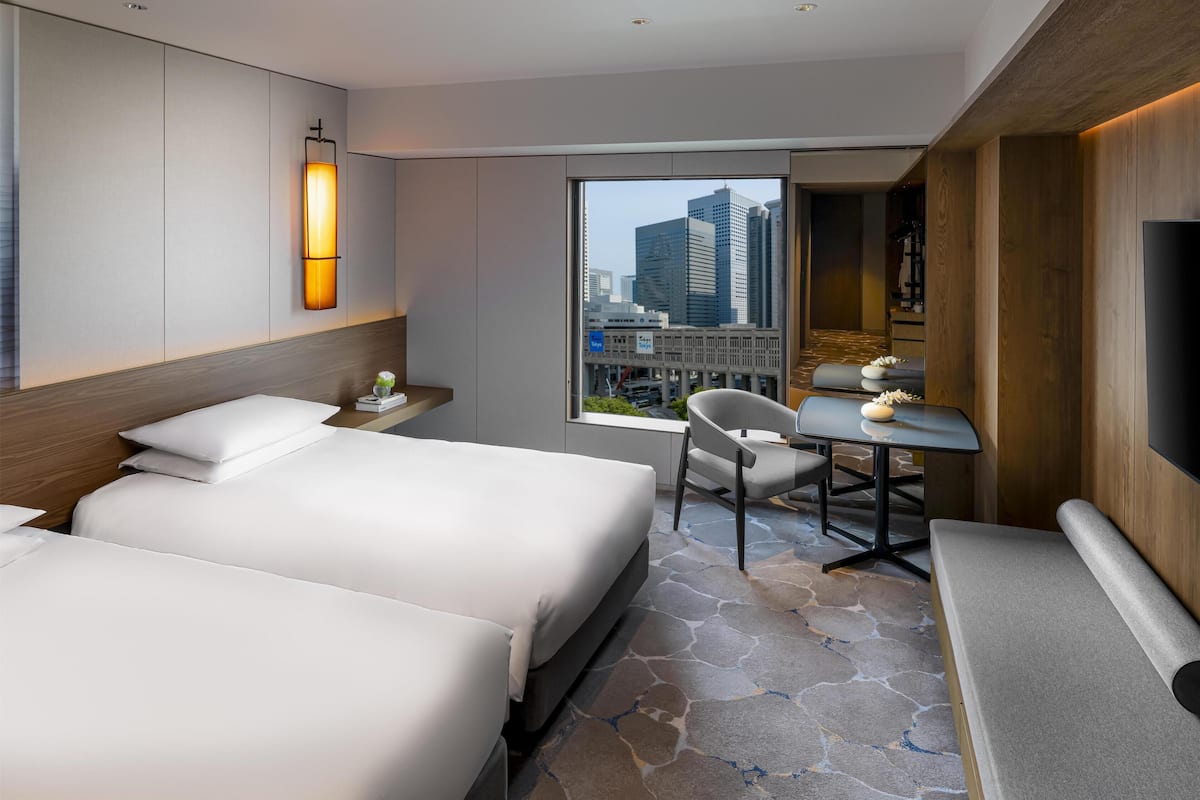 A hotel room with two beds, a desk, a chair, a TV, and a large window with a city view.