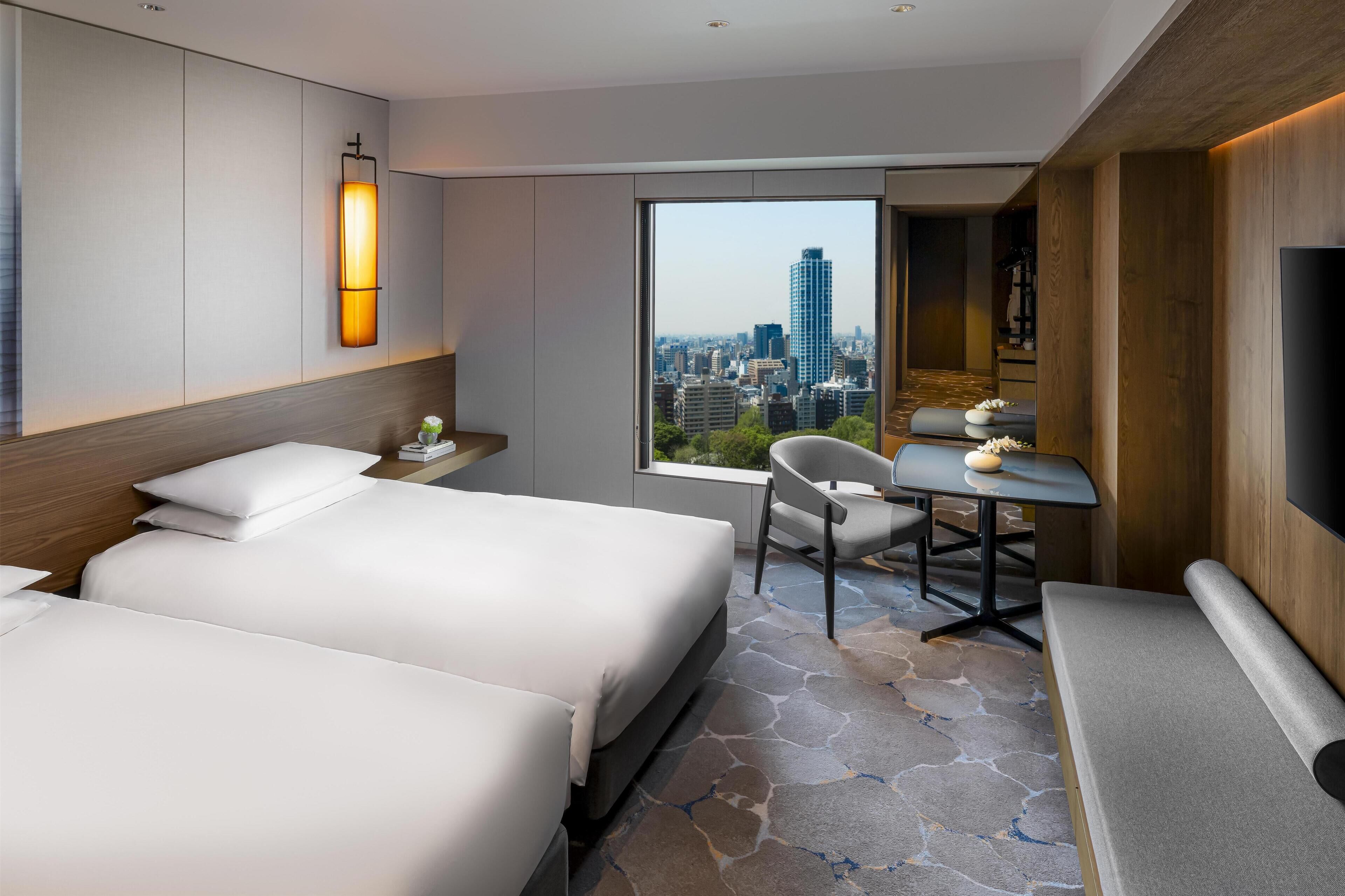A hotel room with two beds, a desk, a chair, a TV, and a large window with a city view.
