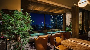 A modern indoor seating area with tables and chairs, large windows with a city view, and a wooden ceiling.