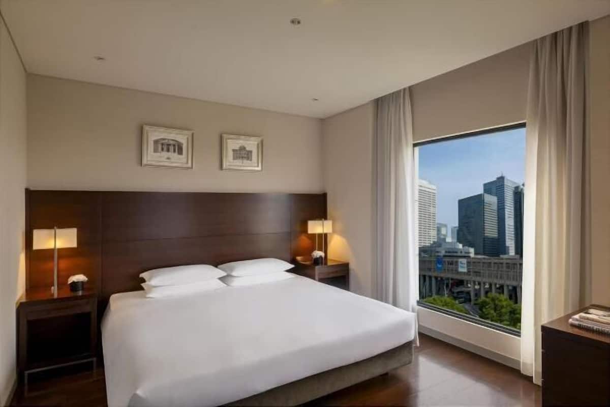 A hotel room with a large bed, two bedside tables with lamps, a view of the cityscape through a large window, and two framed pictures on the wall.