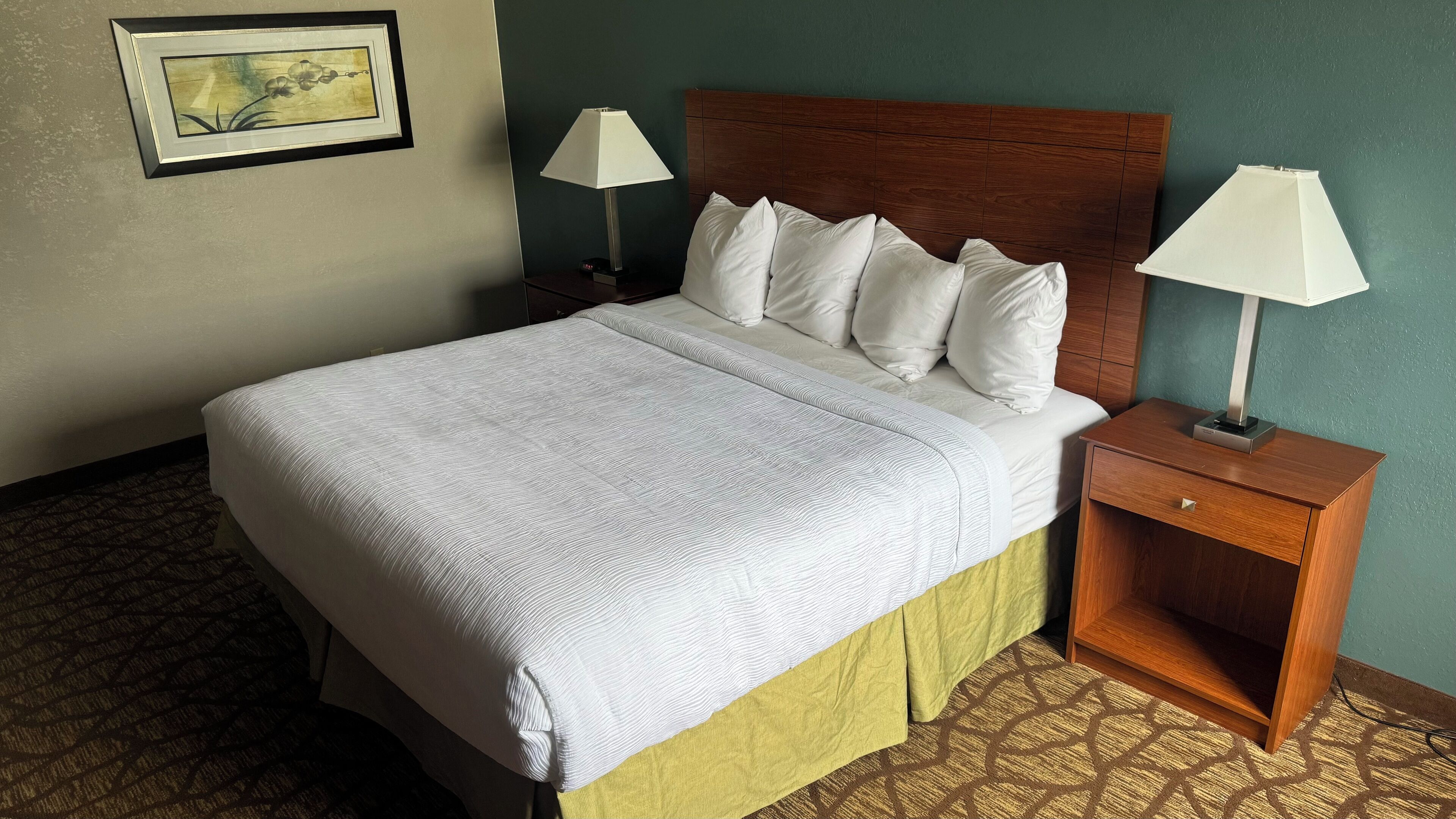 A neatly made bed with a white comforter and pillows, a wooden headboard, two bedside lamps, and a small wooden nightstand with a drawer.