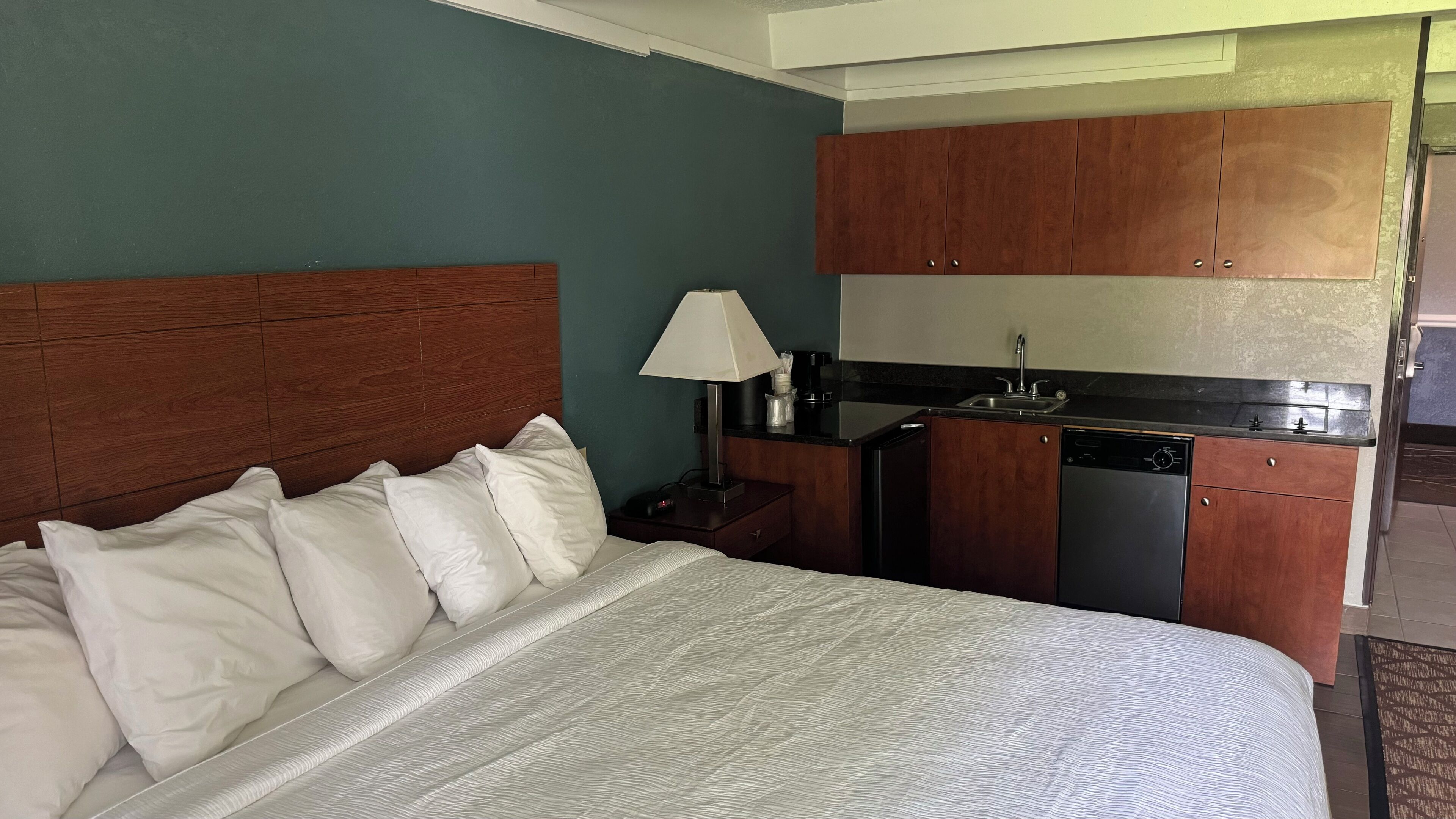 Habitación de hotel con cama, mesita de noche, lámpara, kitchenette pequeña y baño.