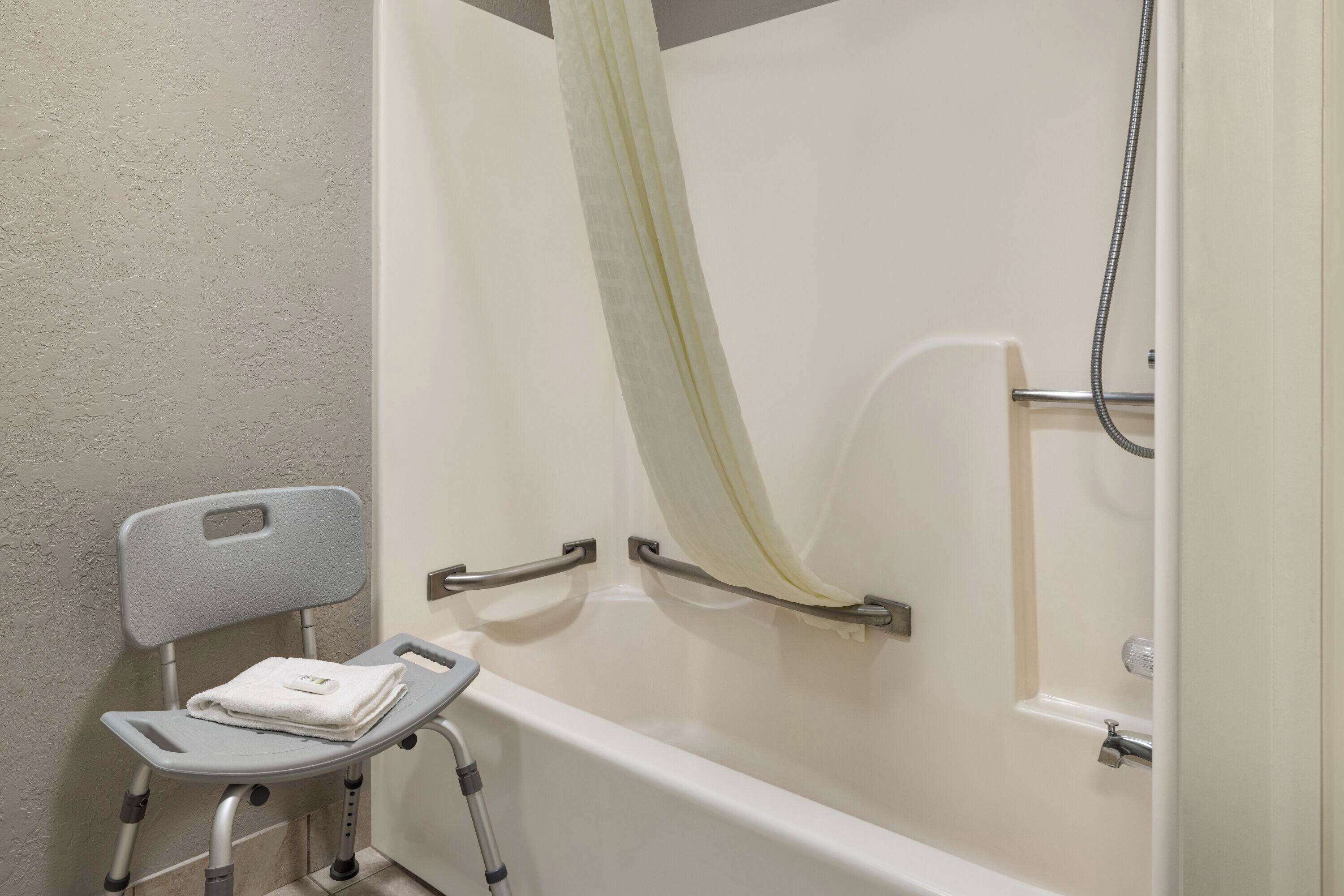A bathroom with a shower, a grab bar, a chair with a towel, and a handheld showerhead.