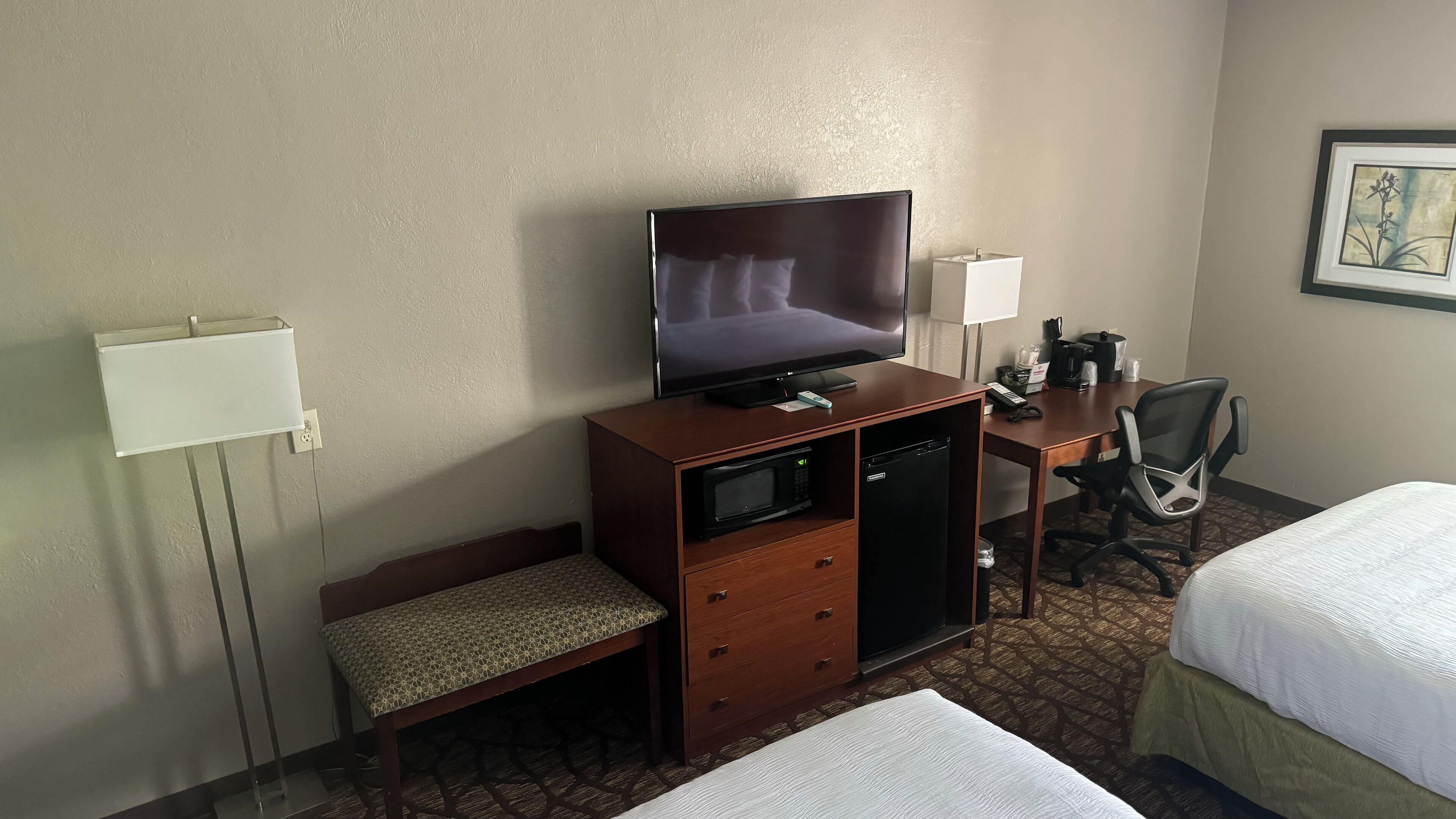 Quarto de hotel com uma cama, uma escrivaninha com televisão, micro-ondas, mini-frigorífico, uma cadeira e uma mesa de cabeceira.