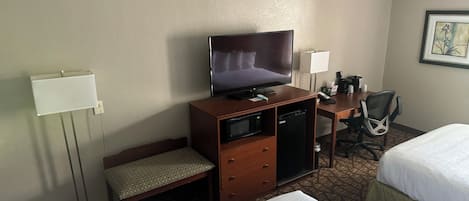 A hotel room with a bed, a desk with a television, a microwave, a small refrigerator, a chair, and a bedside table.