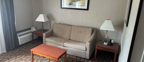 Ein Hotelzimmer mit einer Couch, zwei Nachttischen mit Lampen, einem Couchtisch, einem gerahmten Kunstwerk an der Wand und einem Fenster mit Vorhängen.