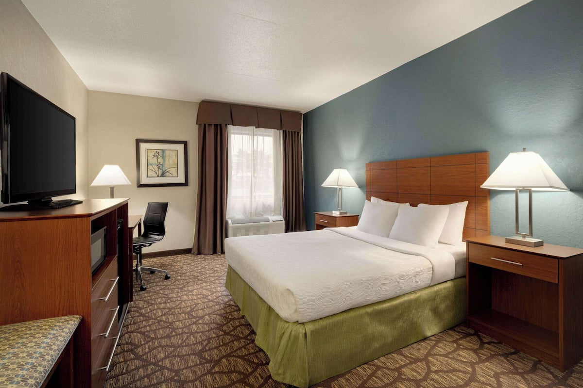 A hotel room with a bed, two bedside tables with lamps, a desk with a television, a chair, and a window with curtains.