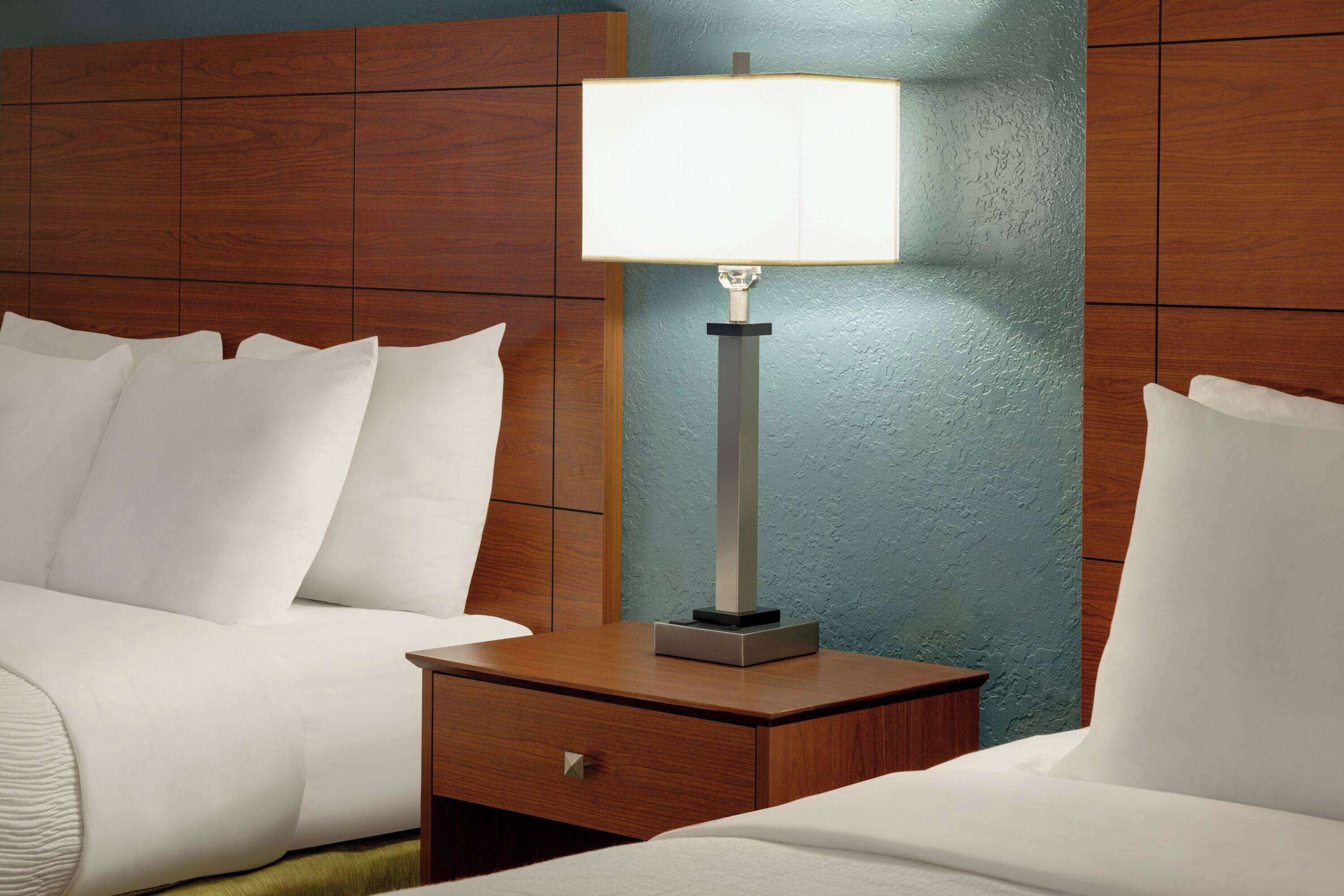 A hotel room with a wooden headboard, a bedside table with a lamp, and a bed with white bedding.
