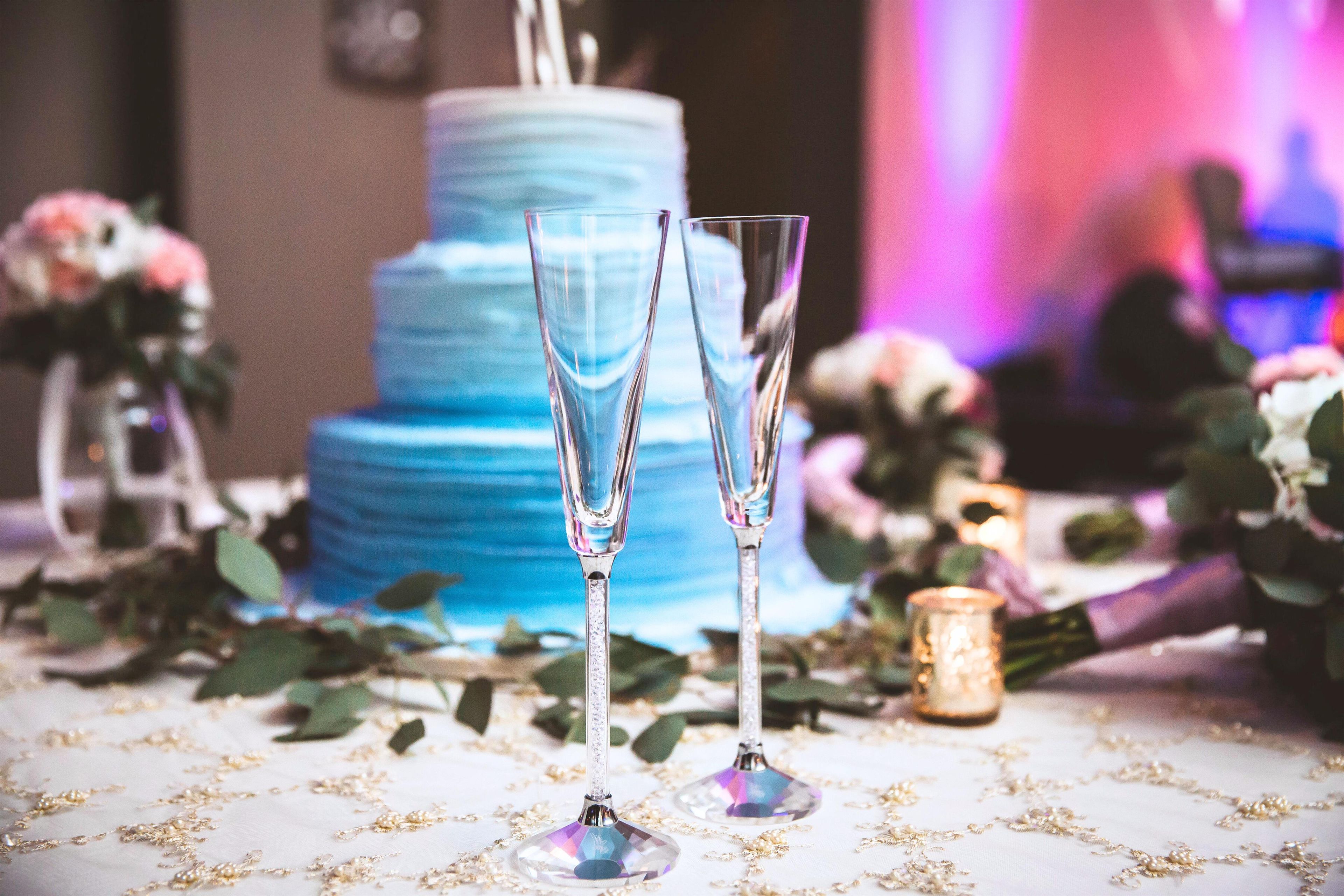 Two champagne flutes on a table with a multi-tiered blue cake in the background.