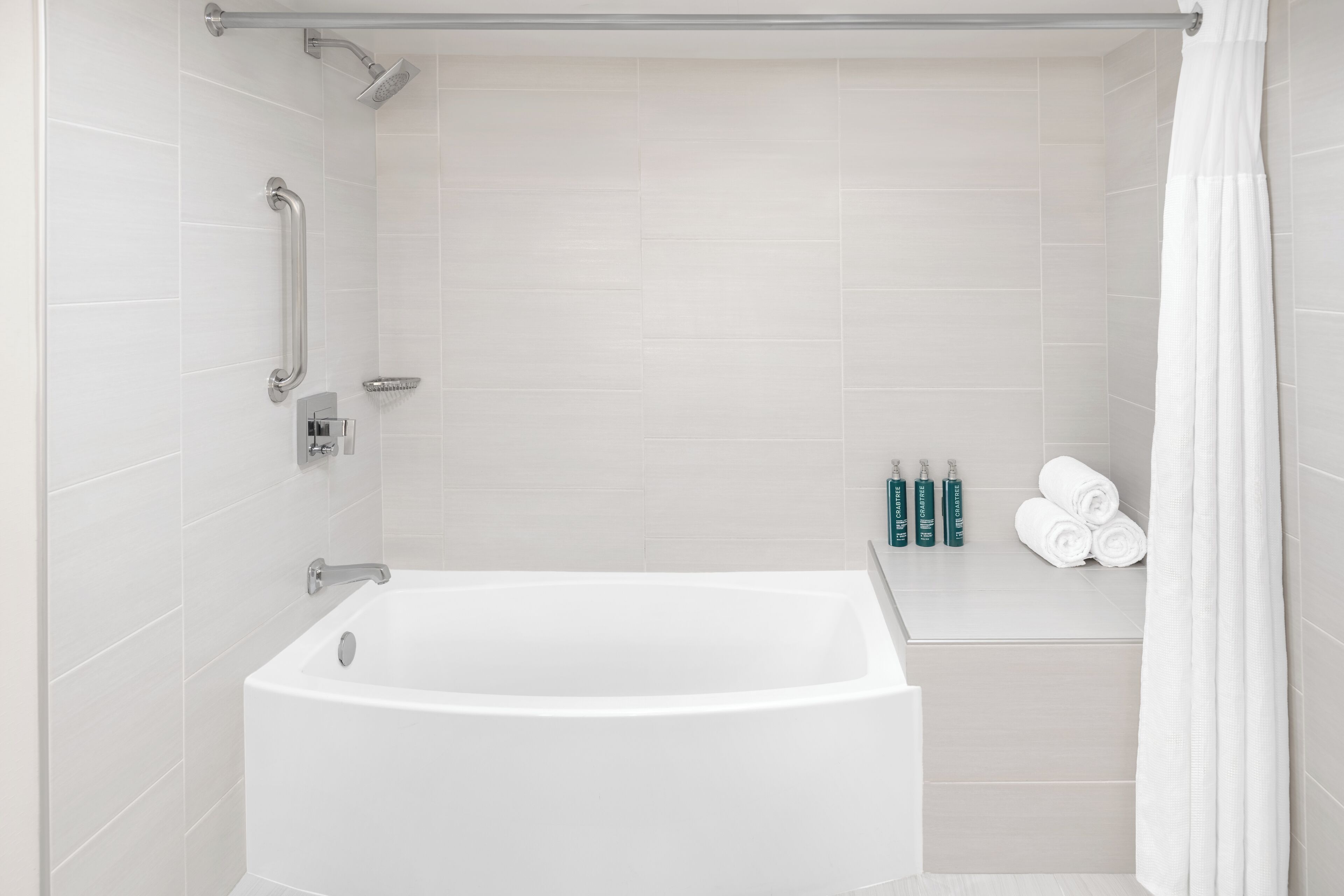 A modern bathroom with a white bathtub, a shower with a handheld showerhead, and a grab bar. There are toiletries on the wall and towels on a shelf.