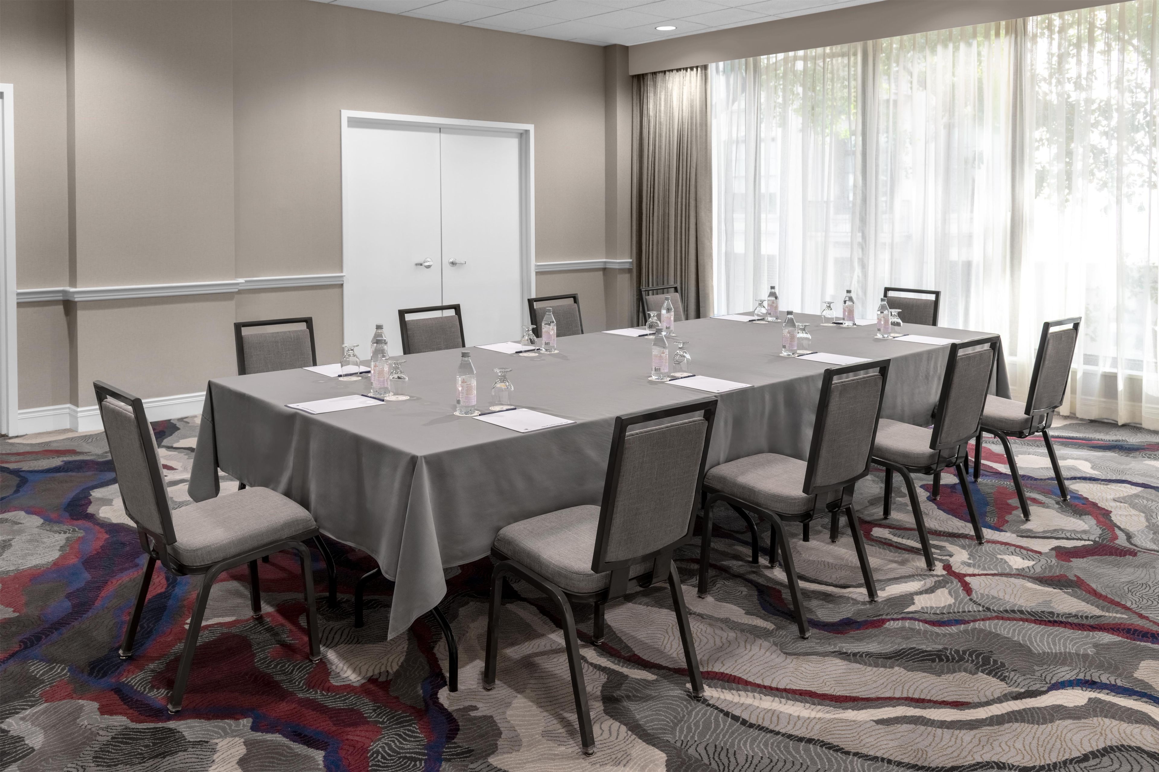 Un salón de conferencias con una larga mesa, sillas, botellas de agua y piso alfombrado.