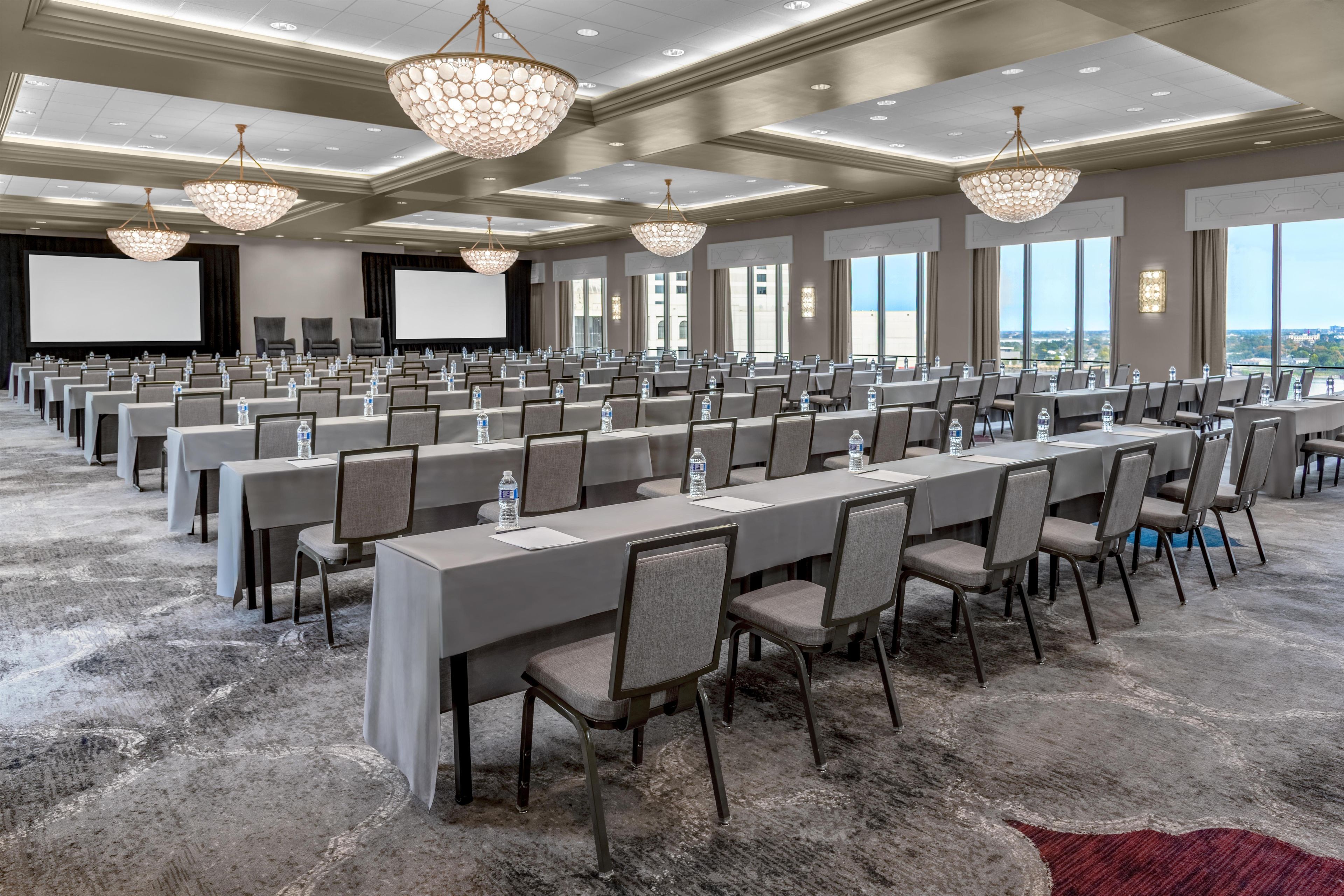 A conference room with rows of tables and chairs, large chandeliers, and a projector screen.