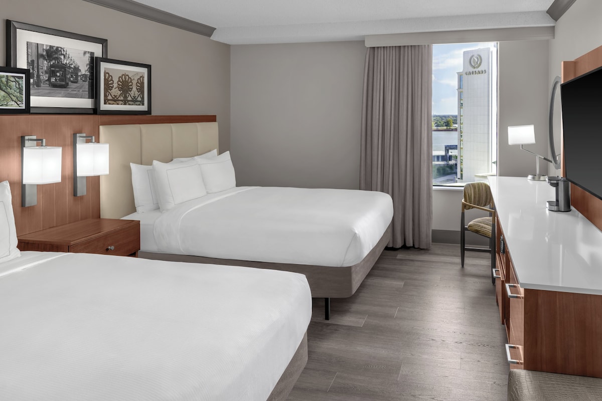 Habitación de hotel con dos camas, un escritorio con televisor y ventana con vista a un edificio.
