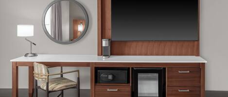 Una habitación de hotel moderna con un escritorio de madera, un espejo redondo, un televisor de pantalla plana, un refrigerador pequeño y una silla.