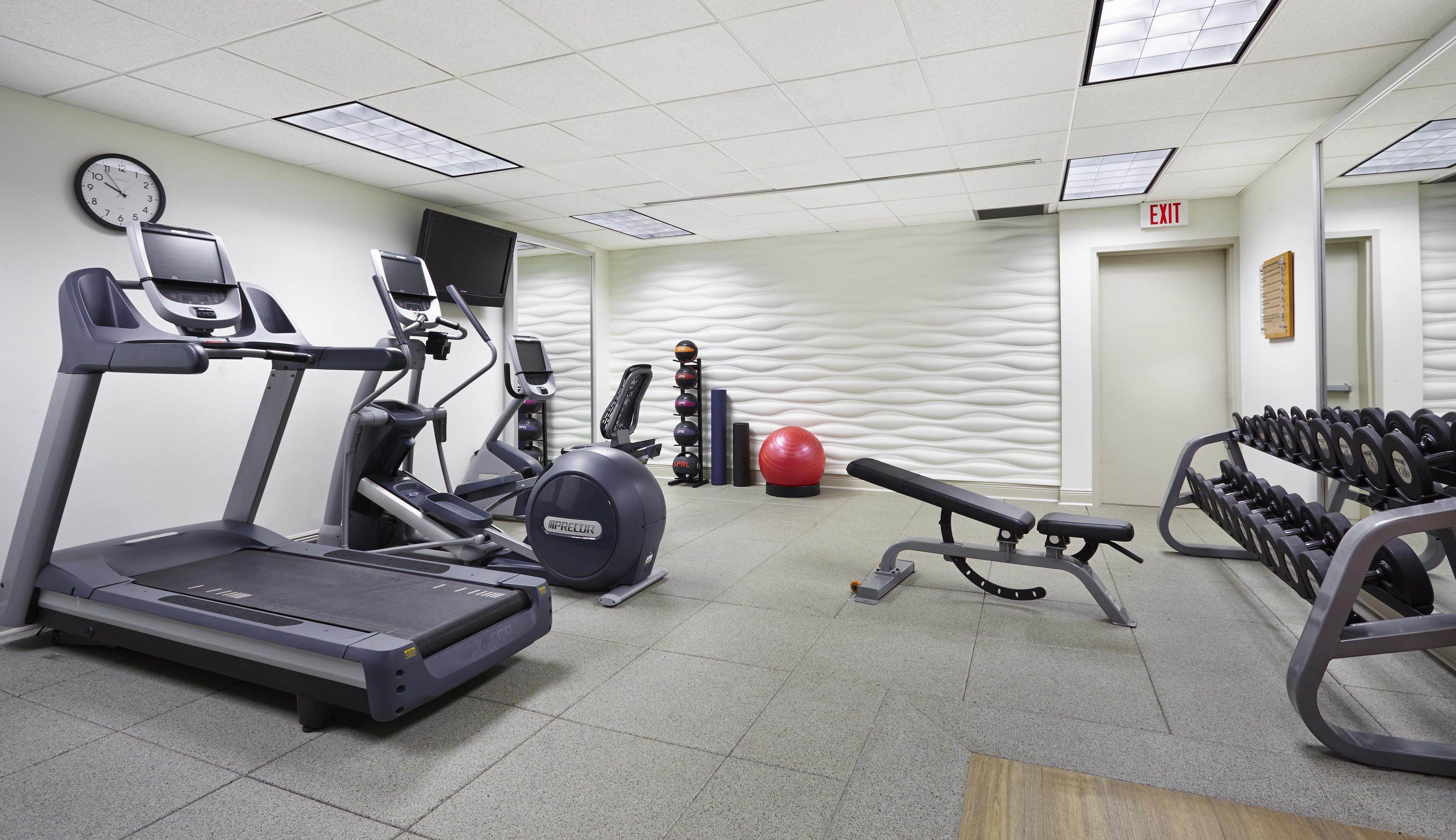A gym with treadmills, an elliptical, a bench press, a set of dumbbells, a red exercise ball, and a wall clock.