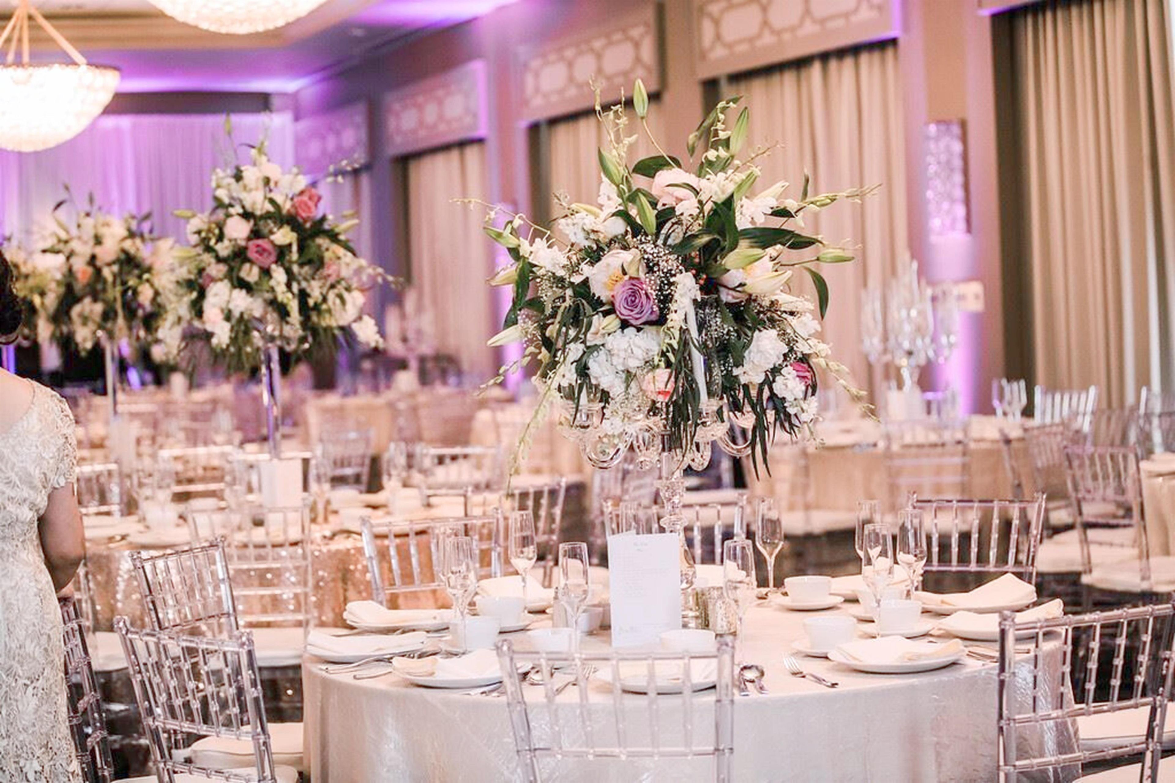 A banquet hall with round tables set for a formal event, featuring floral centerpieces and clear chairs.