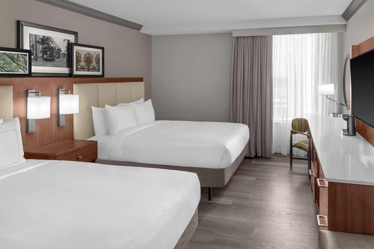 A hotel room with two beds, a desk, a TV, and framed pictures on the wall.