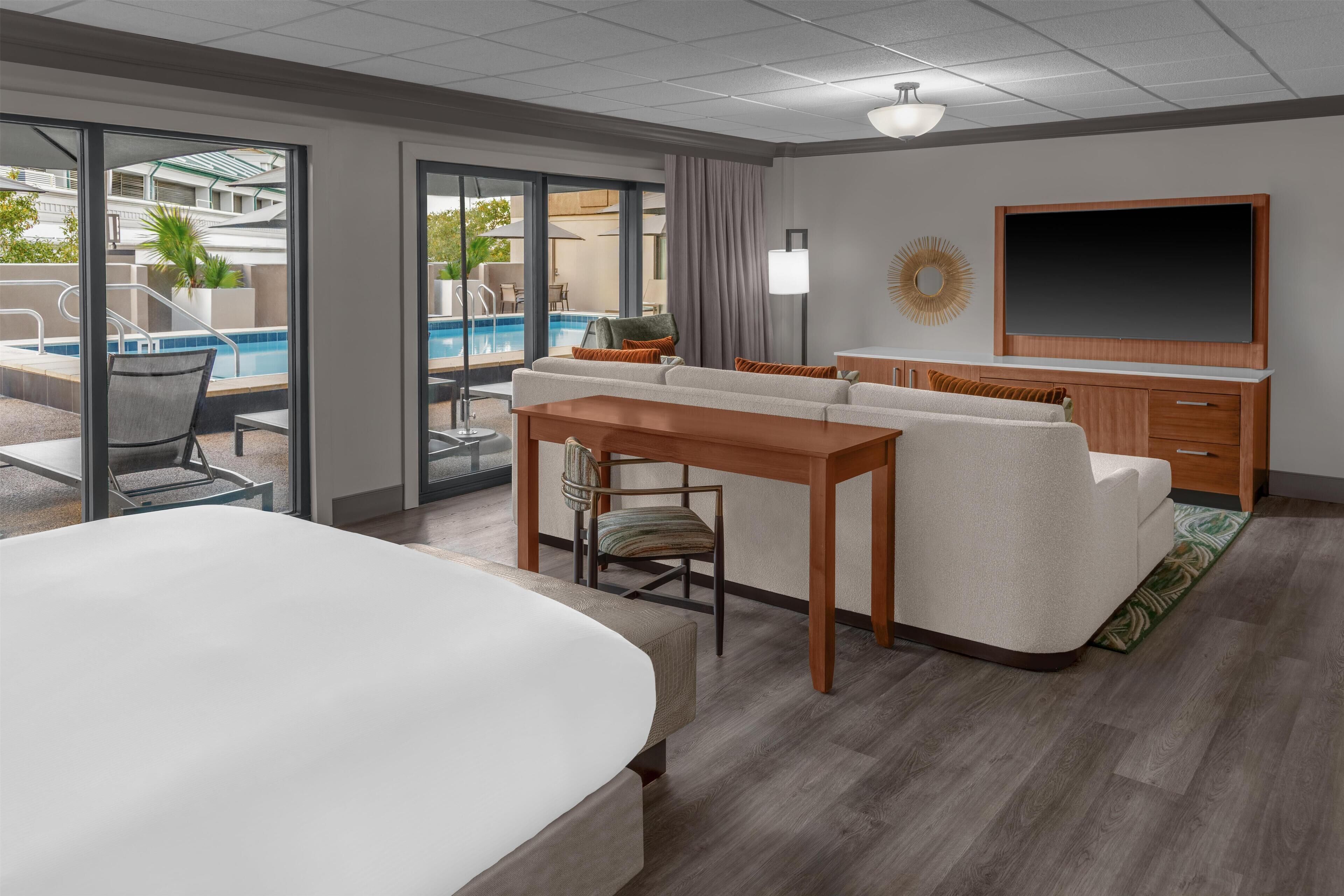 A hotel room with a large bed, a sofa, a dining table, and a TV. There is a pool visible through the glass doors.