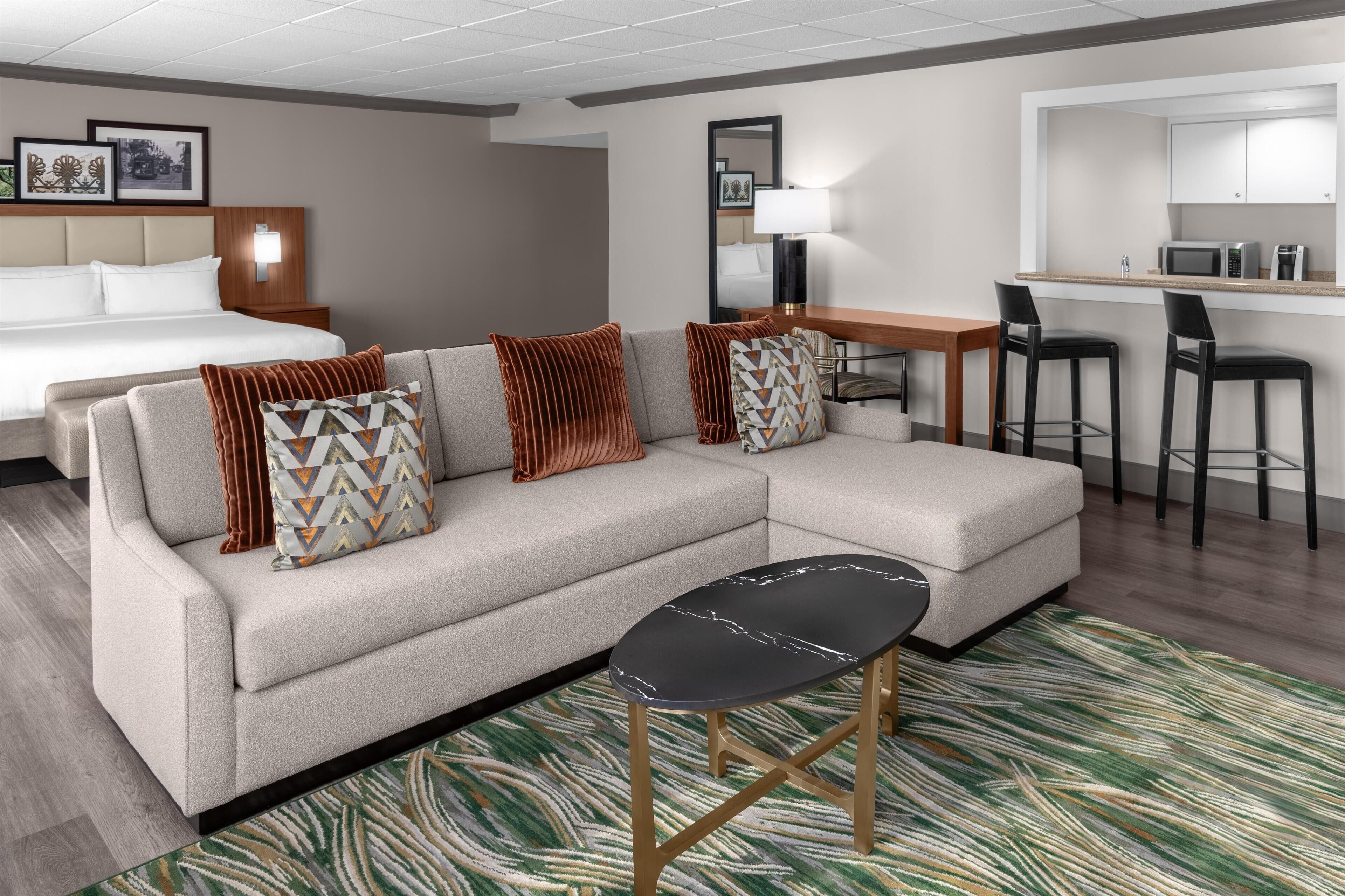 A hotel room with a large bed, a sofa, a small table, a bar area, and a kitchen.